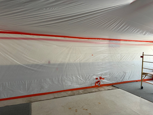 Plastic sheeting with red tape setup for containment during construction or renovation work, with scaffolding visible to the side, creating a sealed barrier in an indoor environment.