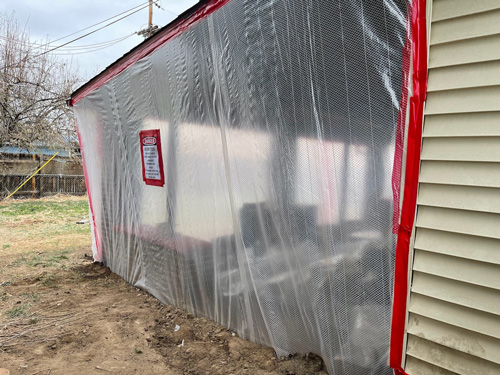 House with an exposed wall covered in clear plastic sheeting, secured with red tape as part of asbestos containment and removal process.